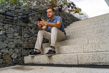 Fototapeta premium Portrait of handsome man sitting on stairs outdoors, using mobile phone and thinking. Business man checking e-mail