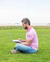 Naklejka na ściany i meble smiling freelance man post blog online sit on grass, networking