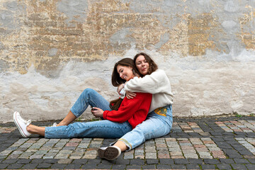 Obraz na płótnie Canvas two young women hugged sitting on a paved sidewalk by an old wall