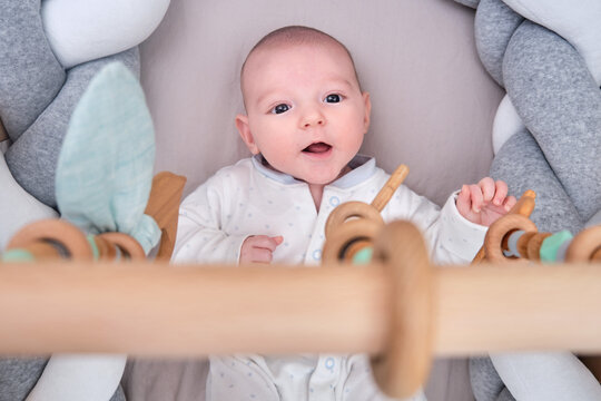 Smiling infant baby boy playing with a wooden mobile suspended above the crib. Happy child playing in bed with toys
