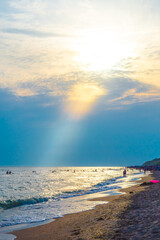 A sunbeam illuminates the sky with clouds in orange and a summer sea beach with tourists.