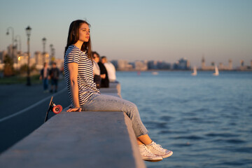 Teen skater girl sit at sidewalk during sunset at seaside. Cute young woman enjoy summer evening at...