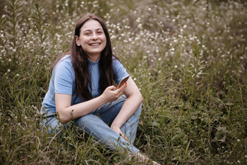 Smiling teenage girl using a phone, sitting on the grass. Beautiful caucasian woman with smartphone outdoors. Happy girl texting on a smart phone in the park. Technology and modern lifestyle concept. 