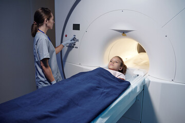 Little girl lying on table of mri scan machine while gloved female radiographer pushing button on...