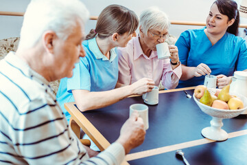 Seniors and caregivers having coffee time in the nursing home, eating and drinking