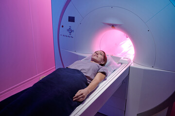 Young female patient lying on long table of mri scan machine while undergoing medical examination - obrazy, fototapety, plakaty