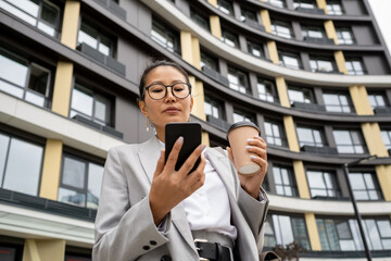 Young businesswoman with drink texting in smartphone or communicating through video chat