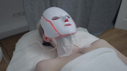 Young Caucasian man lying on beauty bed with red LED light device on face. Relaxed male client enjoying painless effective skin rejuvenating and cleansing procedure in spa indoors