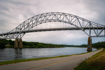 Sagamore Bridge over Cape Cod Canal and bikeways along the riverbank. Dramatic cloudscape over the arching bridge over the river. 