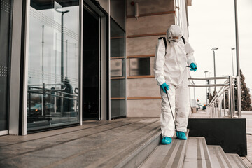 A male person in a white protective suit and mask disinfects the front door and stairs to the office building with chemicals. COVID-19 prevention sanitizing, corona, pandemic