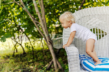 The toddler is playing in the backyard. The baby enjoys childhood, blonde-haired kid in a white bodysuit for children leans on the side of a large linen beige chair. Enjoying the sun, family moments