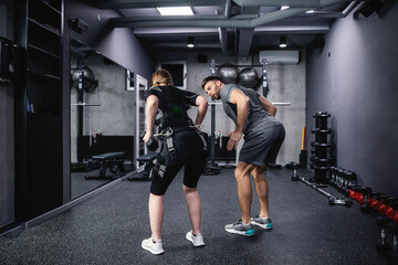 Fototapeta na wymiar EMS training concept. Shot from the back of a female athlete wearing an EMS suit and a male trainer in grey sportswear helping and showing her the exercise with dumbbells Electrical muscle stimulation