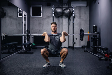 Fototapeta na wymiar Sports routine and active lifestyle. Front view of a young man in gray sportswear squatting with barbell in an indoor gym with equipment. Fitness body fell good and sexy, an attractive sports guy