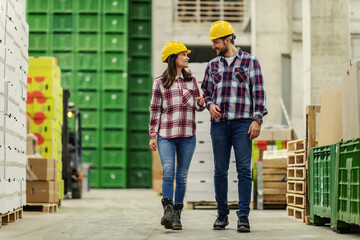 The man and woman with helmet pass through the factory’s wide and modern warehouse in the same plaid clothes. They make plans for the delivery and distribution of goods Management transport logistics