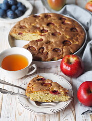 Laid table with freshly baked summer round pie with apples and plums in a white ceramic tin.A cut piece of cake in the foreground. Food background.