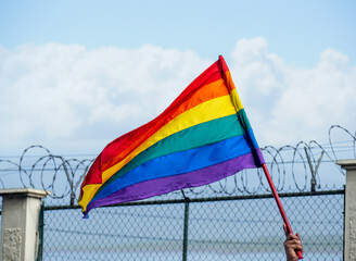 An LGBTQ + flag fluttering in the breeze with a barbed wire fence in the background and a blue sky...