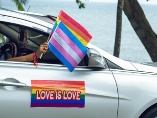 A woman in a car holds two flags, the LGBTQ flag and the Trans Flag, the car has a sticker that says LOVE IS LOVE and has the beach behind