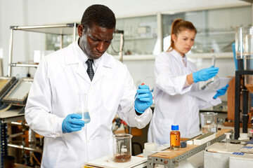 Focused man lab technician in gloves working with reagents and test tubes, woman on background