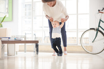 Mother helping baby boy walk in living room