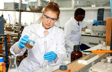 Focused woman lab technicians in glasses working with reagents and test tubes, man on background