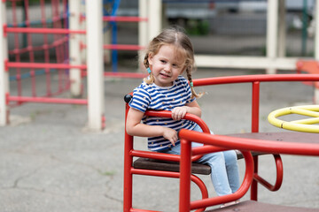 A cute little girl is whirling alone on a red  merry-go-round in a deserted playground.