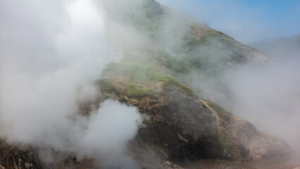Valley of Geysers. Kamchatka. The mountain slopes are shrouded in thick steam from hot springs. Splashes of boiling water are visible. Blue sky in a fog.  