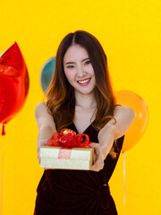 Asian happy pretty girl in sexy dress standing smiling holding green ribbon present gift red box in front colorful star and round shape helium air balloons on yellow background in birthday party