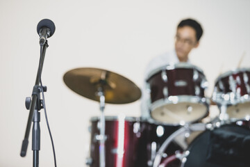 Close-up shot of a black professional microphone on a mic stand with a young male teenage drummer playing the drum in the background. Selective focus at microphone with a musician playing instrument