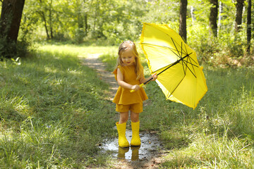 a beautiful blonde girl in a linen suit in yellow boots with a yellow umbrella stands in the rain in the park
