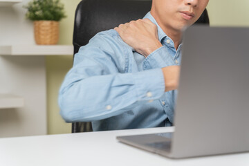 Closeup stressed young businessman massaging stiff shoulder neck when overworked. Exhausted tired office worker using computer laptop suffering from pain, stretching, yoga for pain on shoulder neck.