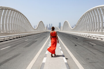 One woman in an orange red dress walking by an empty road on a Meydan bridge with city view on...