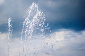 Splashes of a dancing fountain in a summer park against the backdrop of a stormy blue sky. Water jets outdoors in hot weather.