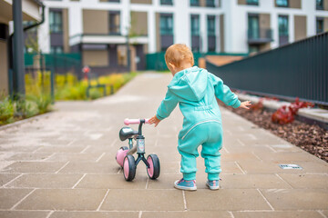 Cute little toddler girl in blue overalls riding on run balance bike. Happy healthy lovely baby child having fun with learning on leaner bicycle. Active kid on cold day outdoors.