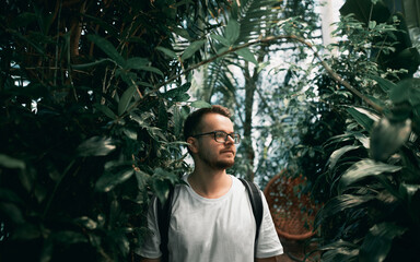 Young freelancer wears glasses in the tropical park. Bearded male in white shirt looks to the side. IT guy wears backpack.