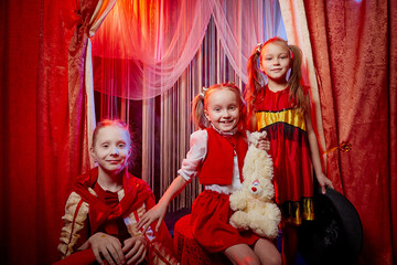 Small girls during a stylized theatrical circus photo shoot in a beautiful red location. Young models posing on stage with curtain. Sisters or female friends together. Twin sisters and a teenager