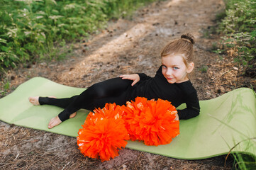 A tired, smiling girl, a child cheerleader in a black suit lies, resting on a green rug in the...