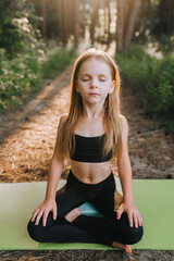 A beautiful blonde preschool girl, a child athlete professional in a black suit sits on a green rug in a lotus position and meditates, relaxes, training yoga exercises in the forest.
