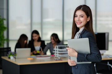 Portrait of young attractive Asian female office worker in formal business suits  smiling at camera in office with blurry colleagues sitting in office as background