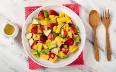 Mixed fruits salad including strawberry, kiwi, apple, and pineapple in white dish place on sackcloth on wooded table.  Utensil spoon and fork and oil salad dressing cup beside
