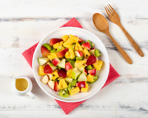 Mixed fruits salad including strawberry, kiwi, apple, and pineapple in white dish place on sackcloth on wooded table.  Utensil spoon and fork and oil salad dressing cup beside