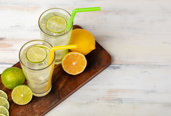 Refreshing glasses of cold fruit juice, ice lime and lemon juice, decorating with fresh lime and lemon slice on wooden chopping board on white table. Yellow and green straw are next to it