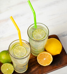 Fototapeta premium Refreshing glasses of cold fruit juice, ice lime and lemon juice, decorating with fresh lime and lemon slice on wooden chopping board on white table. Yellow and green straw are next to it