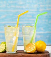 Refreshing glasses of cold fruit juice, ice lime juice, decorated using  fresh lime and lemon slice with yellow and green straw on wooden chopping board on white table in front of blue background