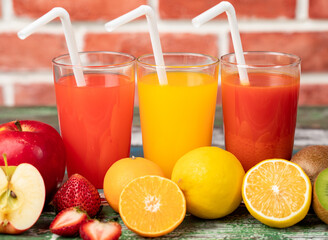 Different fruits juice in glass, apple, orange and strawberry juice with straw, looking refreshing on colourful wood board in front of brick wall background. Picture decorating with fresh fruits