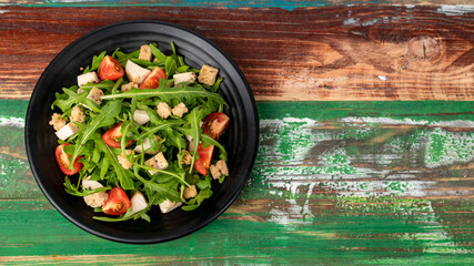 Fresh healthy rocket salad with tomato, crouton, small pieces of herb roasted chicken in black ceramic dish on colourful wooded table. Concept for healthy