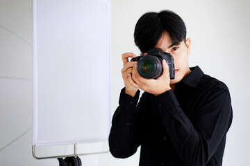 Asian handsome male photographer wearing casual black shirt, holding a camera, posing and sitting in a studio with light equipments.