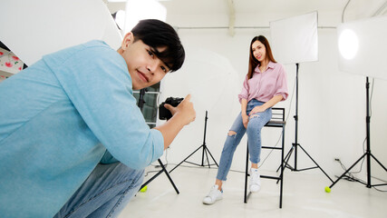 An asian handsome male photographer taking photo of female model wearing casual shirt with jeans, sitting and posing in indoor studio with blur background of lighting equipments.