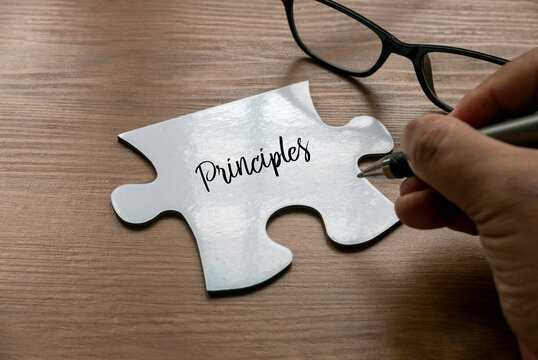 Selective focus of a hand writing word ' Principles ' on a a piece of white jigsaw puzzle with a glasses on wooden background.