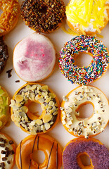 Top view flat lay of a colorful group of twelve delicious donuts in a white paper box on a wooden table. Collection of dozen donuts with glazes, sprinkles, almonds, peanuts, and chocolate chips