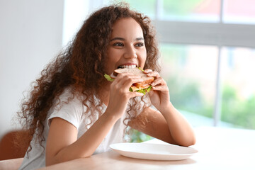 Young African-American woman eating tasty sandwich in kitchen